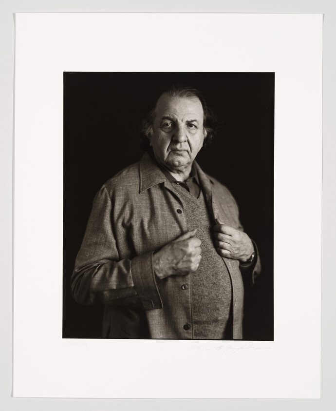 A black and white portrait of an older man with a solemn expression, wearing a buttoned-up jacket over a shirt. He is standing against a dark background and is holding his jacket open slightly with his hands. The photo is framed with a white border, and there is a signature at the bottom right corner.