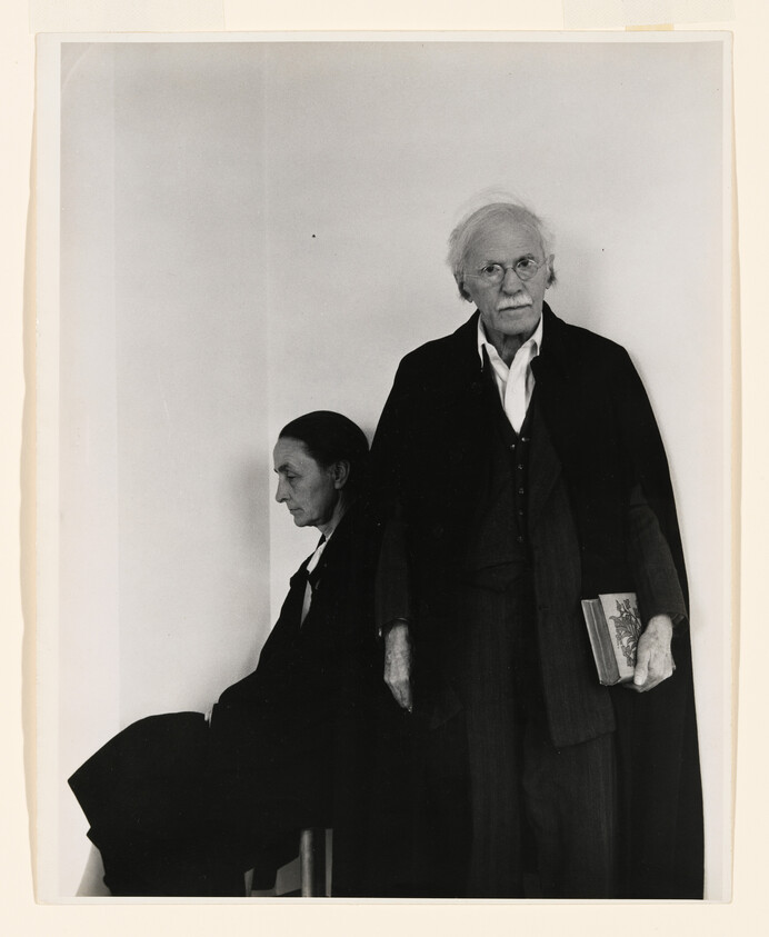 An elderly man stands holding a book while a woman sits turned away against a plain wall.