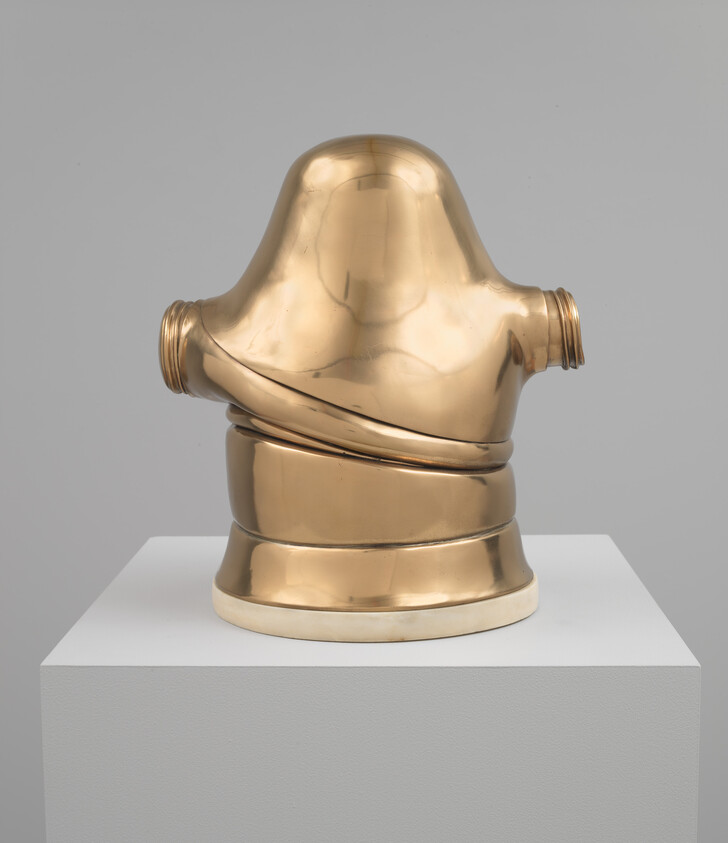 A polished brass helmet-shaped sculpture with threaded side openings sits on a white pedestal.