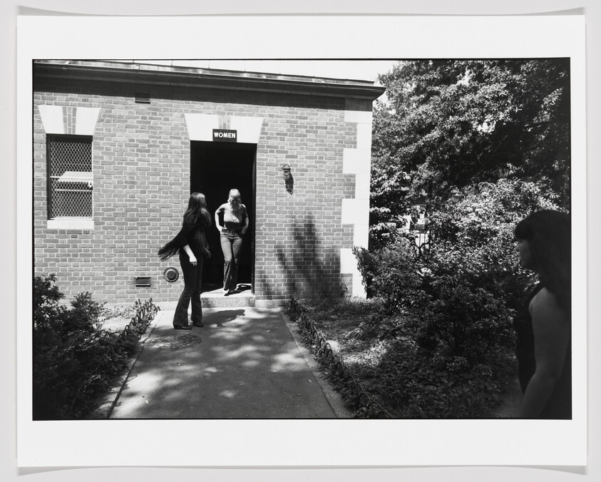 Three women near a brick building as one exits a doorway marked "WOMEN" onto a shaded path.