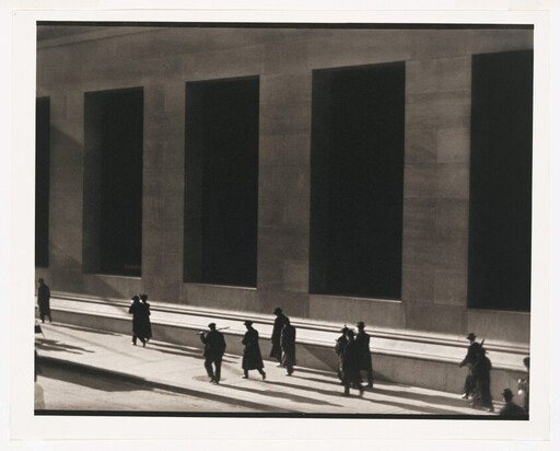 People walk along a sunlit city sidewalk beside a building with tall dark windows.