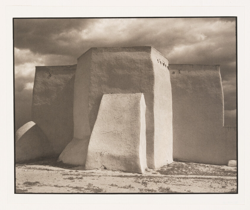 A sunlit adobe building with thick buttresses stands against a dramatic cloudy sky.