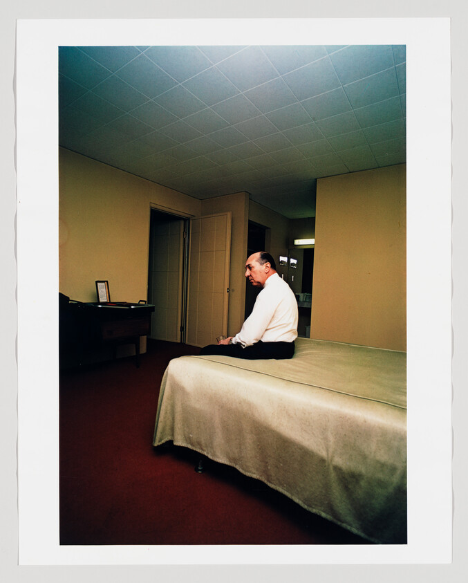 A man in a white shirt sits on the edge of a bed in a sparsely furnished room.