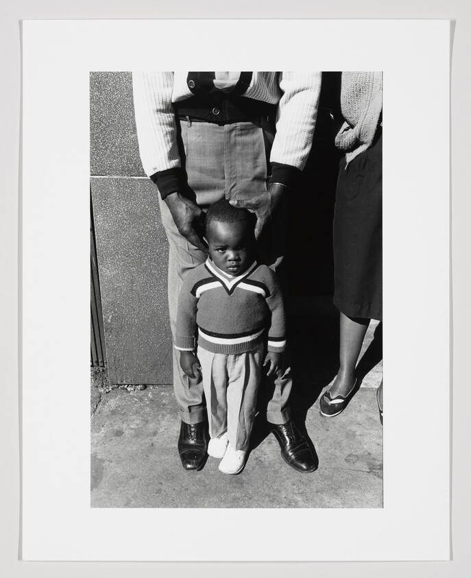 A black and white photograph capturing a young child standing between two adults, whose faces are cropped out of the frame. The child looks directly at the camera with a neutral expression, wearing a striped sweater and pants. The adults' hands rest gently on the child's shoulders, with one adult wearing a white cardigan and high-waisted trousers, and the other in a dark skirt and sweater. The trio is standing on a concrete surface against a wall with a vertical shadow line.