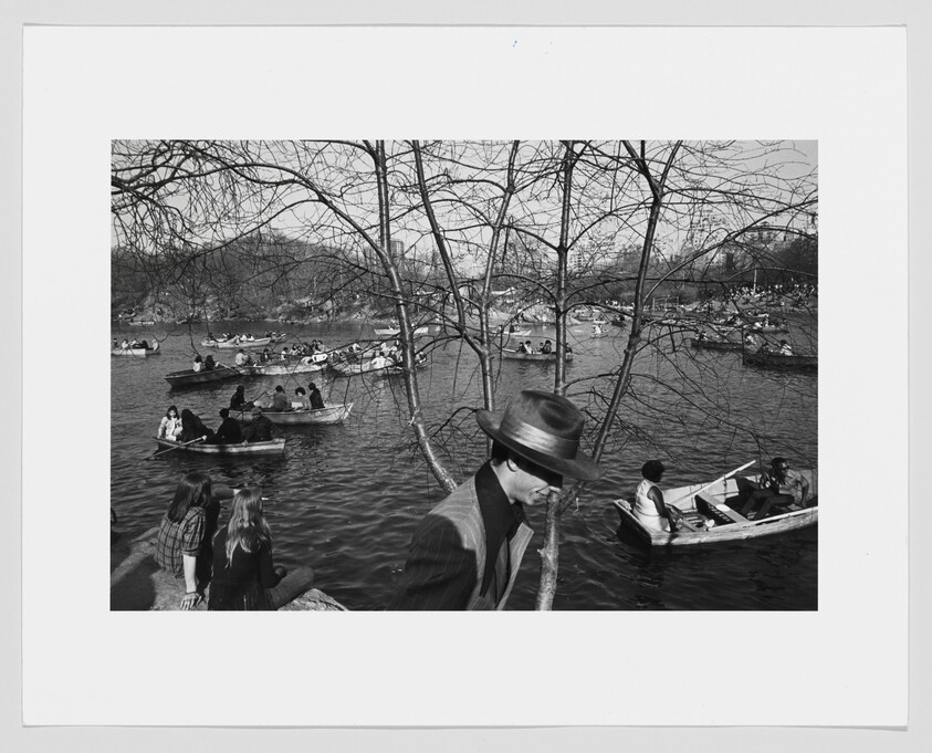 Man in a hat walks past a tree while many people row boats on a busy lake.