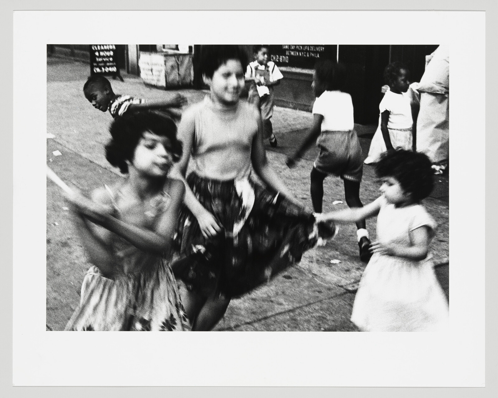 Several children play and spin together on a city sidewalk, one girl tugging another's skirt.