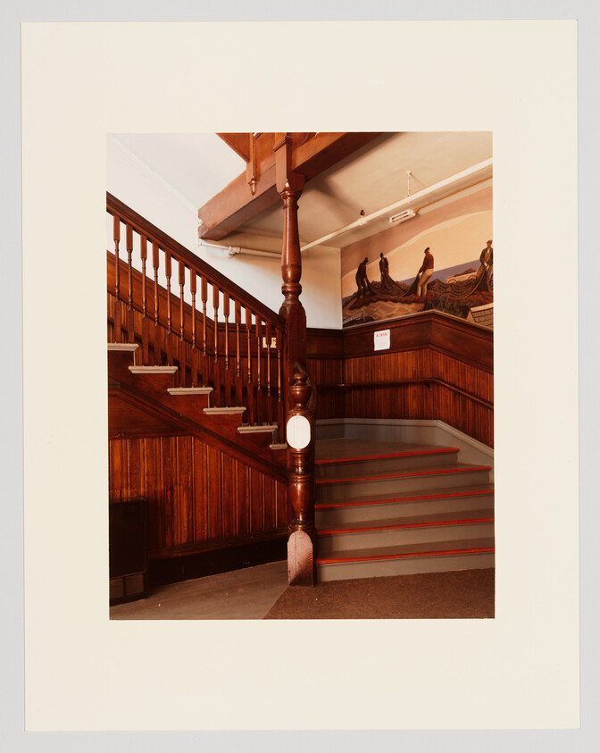 Wide wooden staircase with carved newel post and mural of figures on the upper landing.