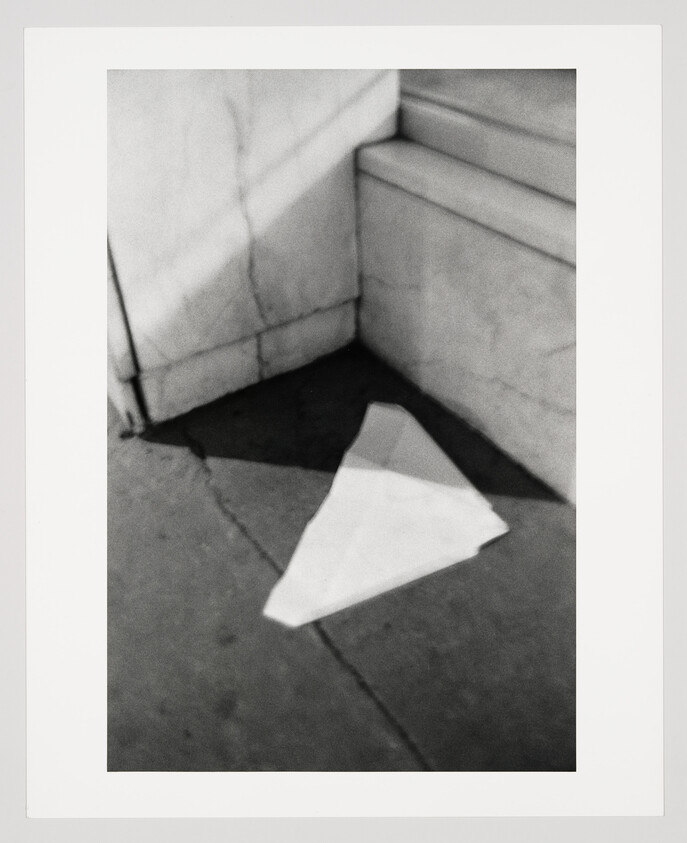 A crumpled paper airplane lies on a cracked sidewalk corner, casting a dark shadow.