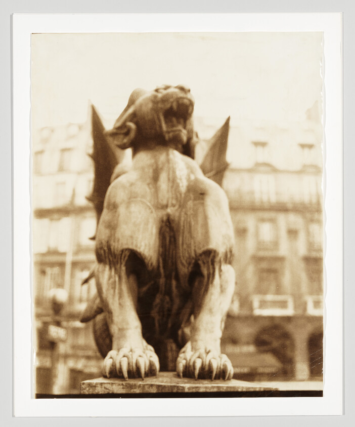 A winged stone gargoyle crouches with open mouth and large front paws on a ledge.