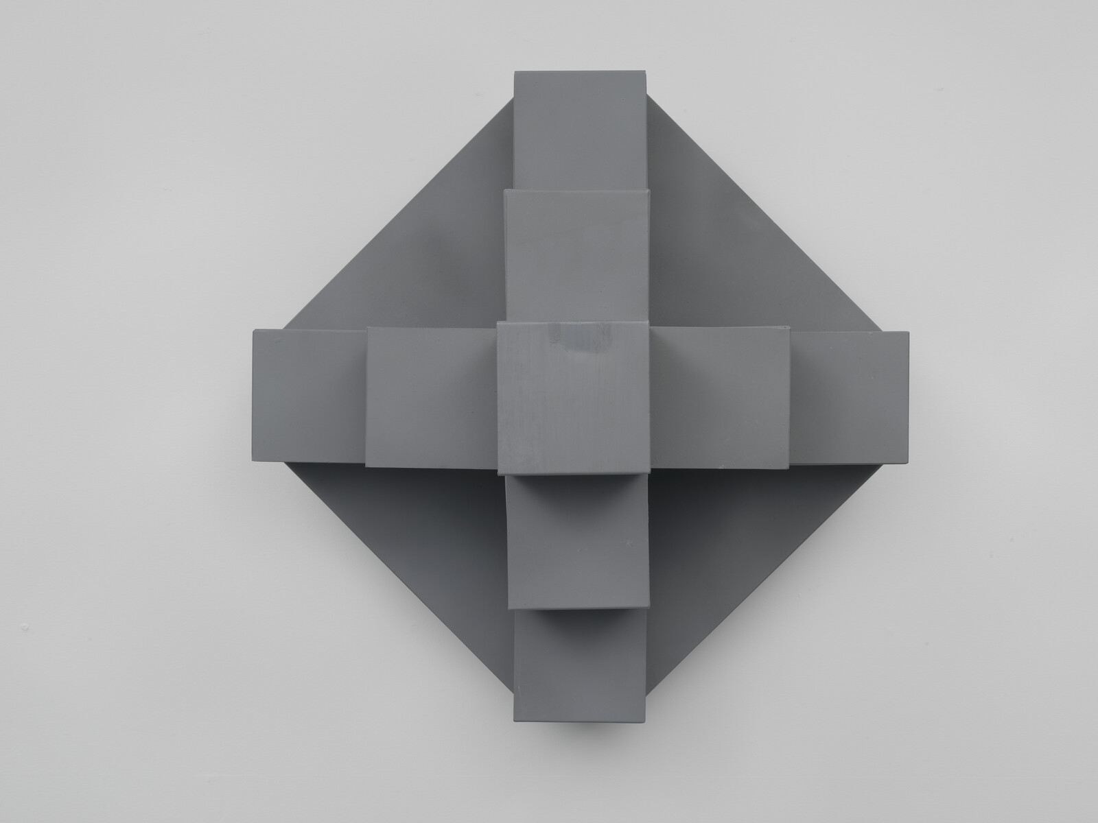 Gray geometric wall sculpture made of stacked rectangular blocks forming a cross over a diamond.