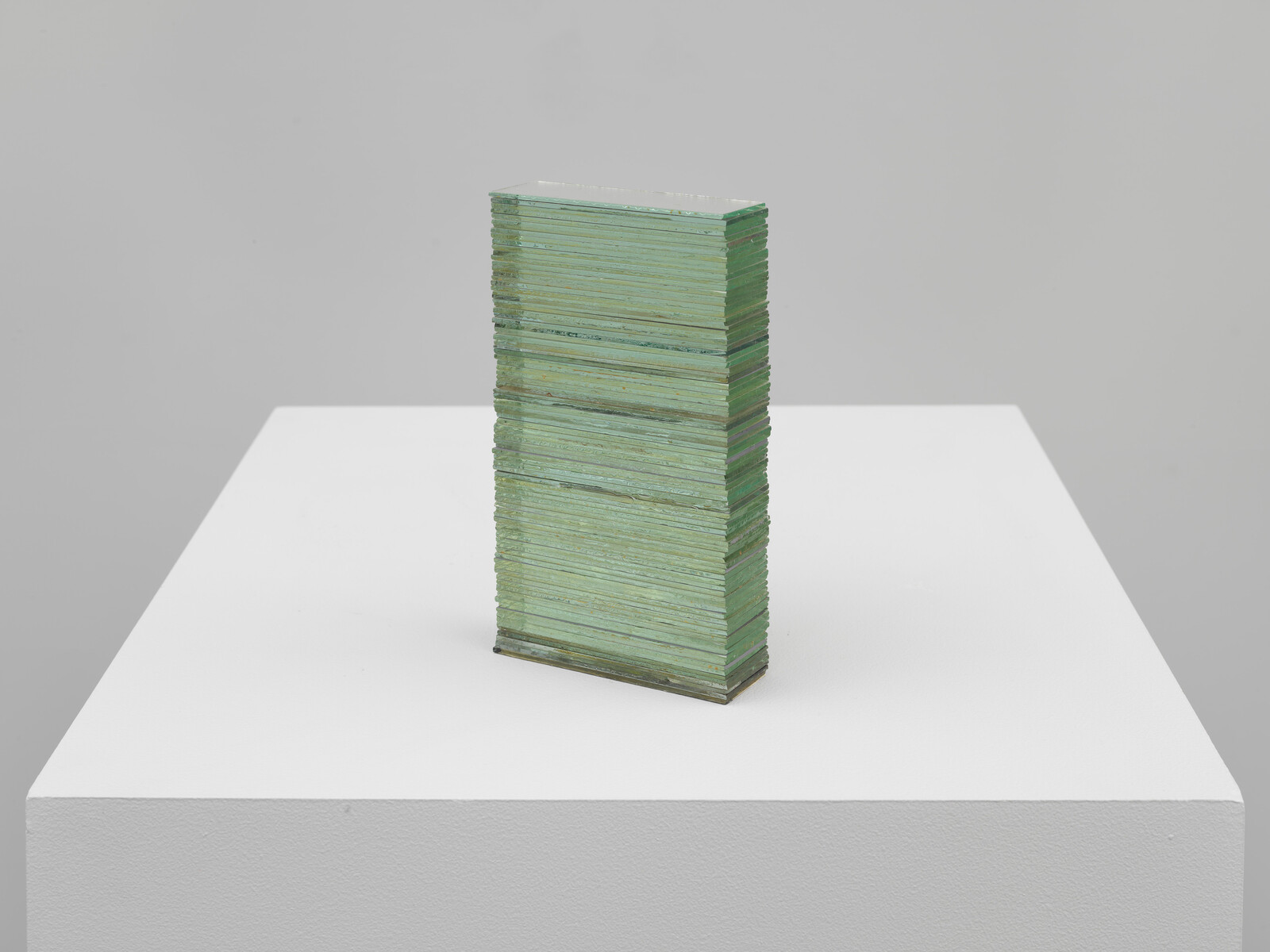 A tall stack of thin green glass panes stands upright on a white gallery pedestal.