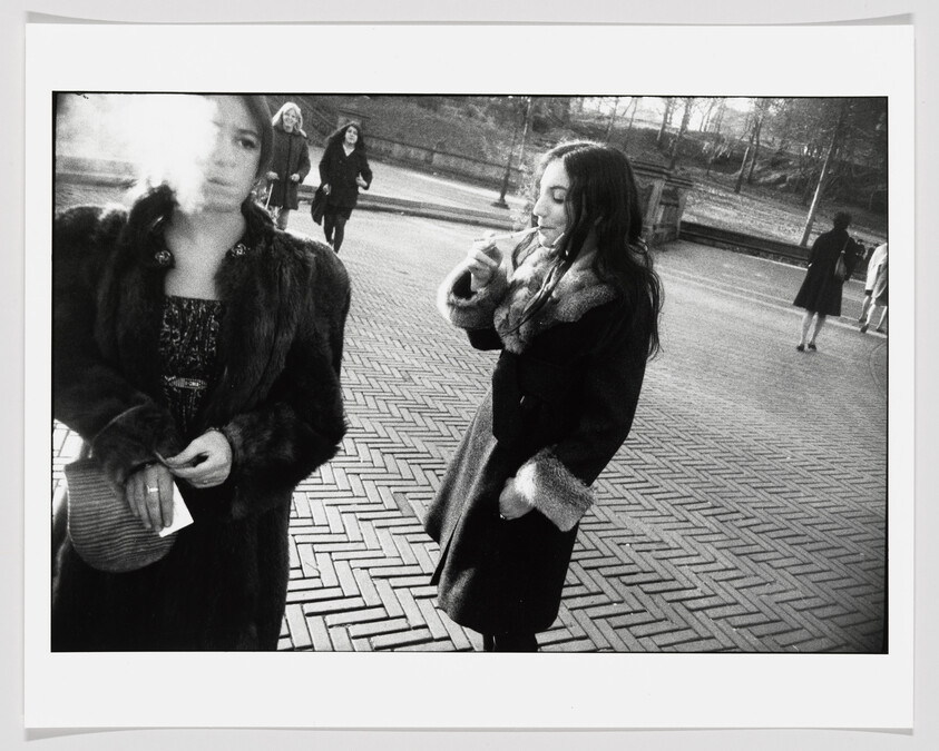Two women in coats stand on patterned pavement, one smoking and exhaling a cloud of smoke.