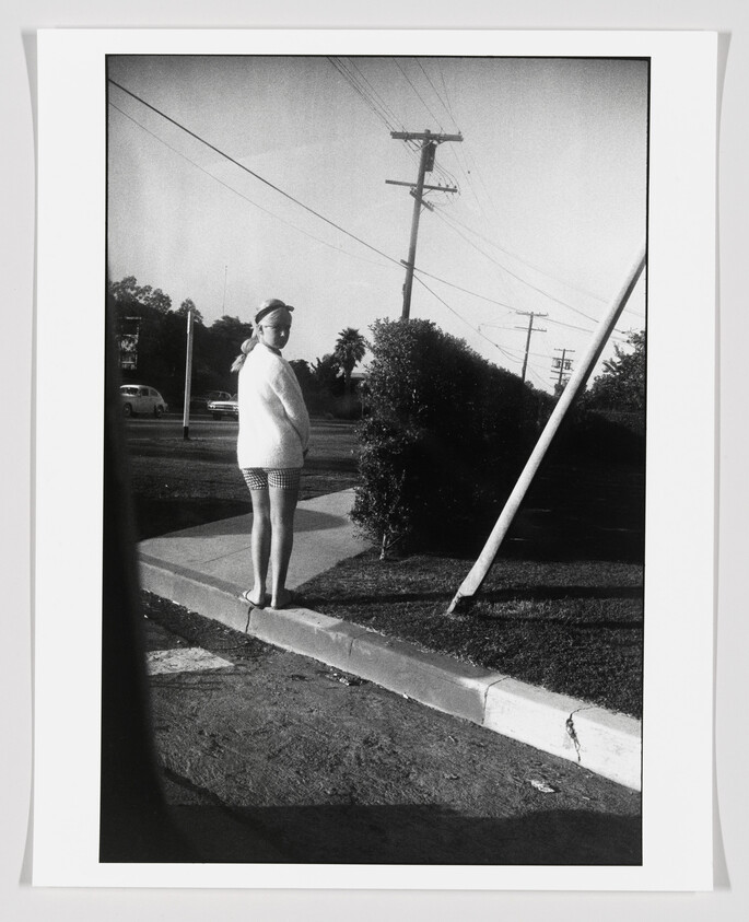 A person stands on a curb looking back toward the camera beside a leaning utility pole.