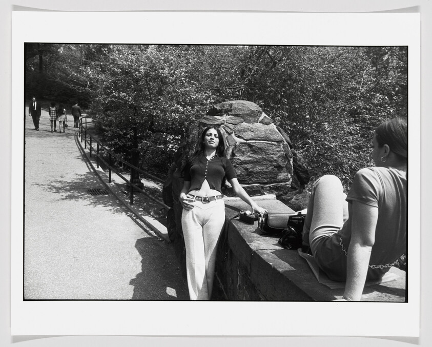 Young woman leans against a stone wall in a park while a friend sits nearby.