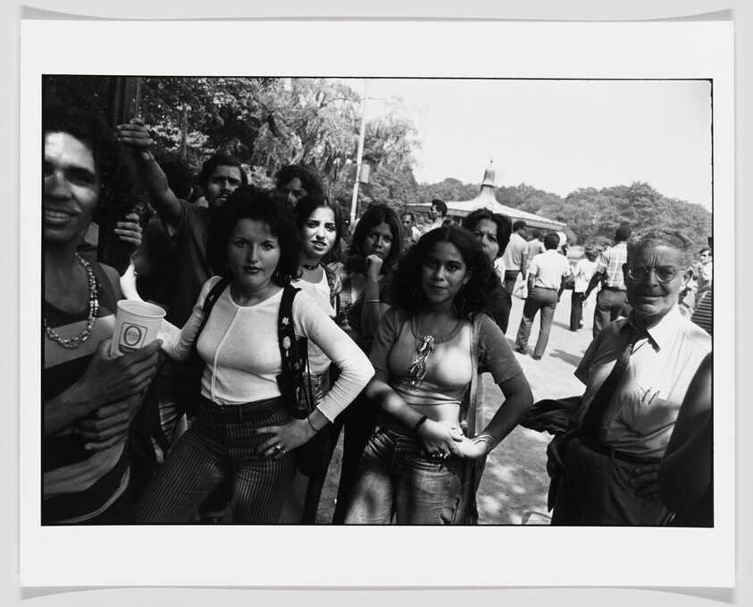 A group of young people pose confidently at an outdoor gathering while others mill about.