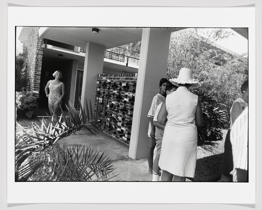 Woman in a swimsuit stands in a doorway while others talk near a house entrance.