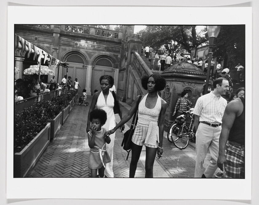 A woman walks hand-in-hand with a young girl while another woman follows in a busy park.