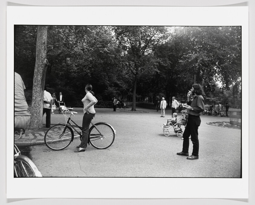 Woman standing with her bicycle in a park while a man nearby smokes and pushes a stroller.