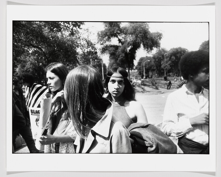 Young people gathered outdoors, a woman with a headband looks directly at the camera while others talk.