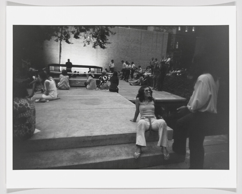 Young woman sits on steps laughing and chatting with a standing friend while others gather nearby.