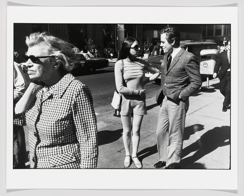 A young woman in shorts talks to a man in a suit on a city sidewalk.