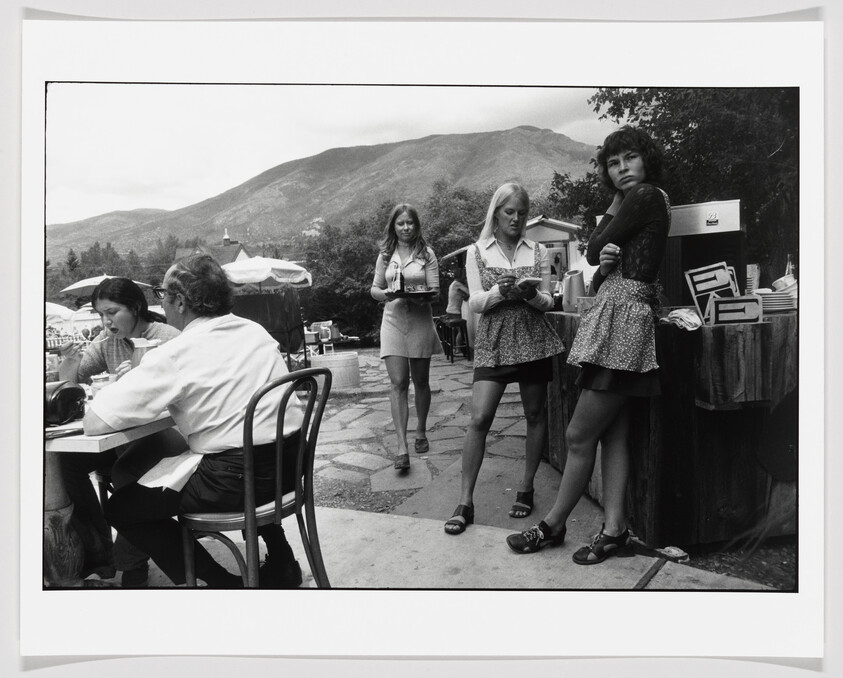 Three young women servers stand and walk at an outdoor café with mountains behind them.