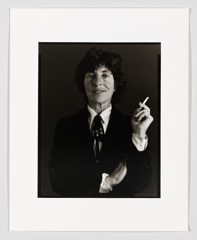 Black and white portrait of a person with short curly hair, wearing a dark blazer, white shirt, and patterned tie, holding a cigarette in their right hand. The person is looking directly at the camera with a slight smile. The photograph is framed with a white border that includes a signature at the bottom right corner.