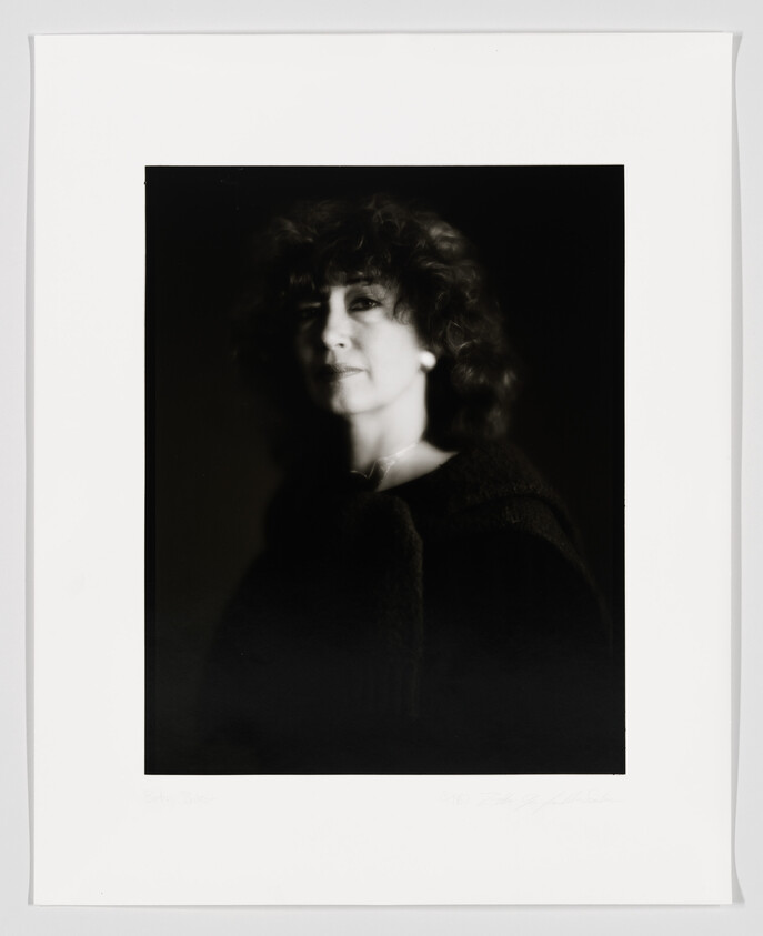 A black and white portrait of a woman with curly hair, looking to the side with a soft focus effect, creating a dreamy atmosphere. The image is framed with a white border, and there is handwriting at the bottom.
