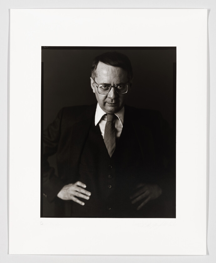 A black and white portrait of a bespectacled man in a suit and tie, looking directly at the camera with a serious expression. The photograph is mounted on white paper with a signature at the bottom.