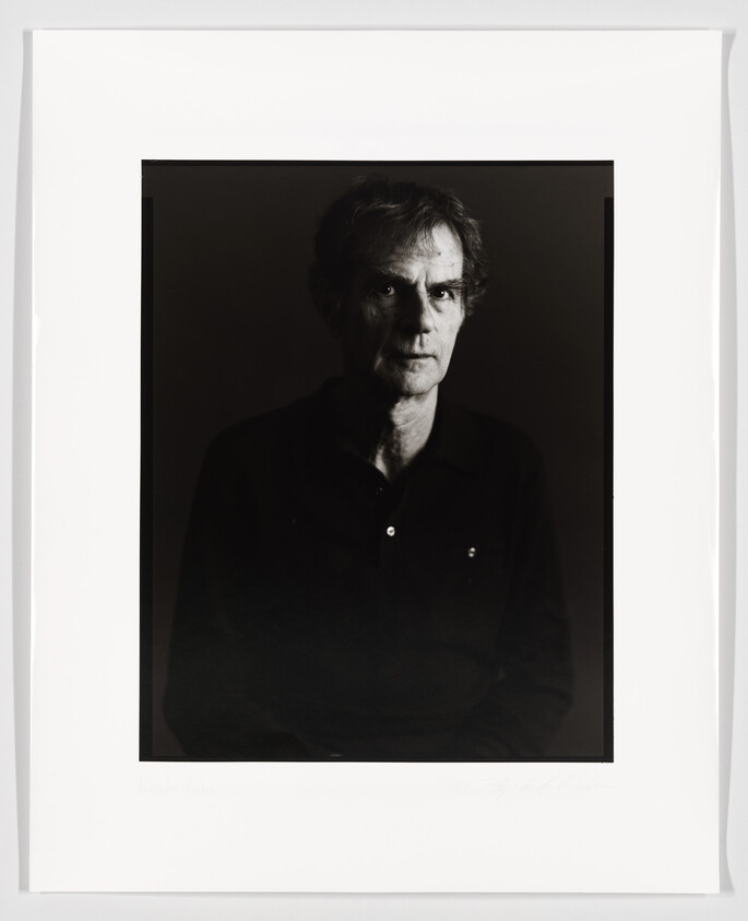 A black and white portrait of a man with a contemplative expression, looking slightly to his left, against a dark background. The photo is framed with a wide white border, and there is handwriting at the bottom.