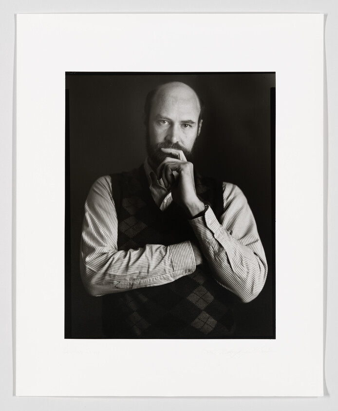 A black and white portrait of a bald man with a beard, wearing a patterned vest over a striped shirt, resting his chin on his hand in a thoughtful pose. The image is framed with a white border that includes a signature and text at the bottom.