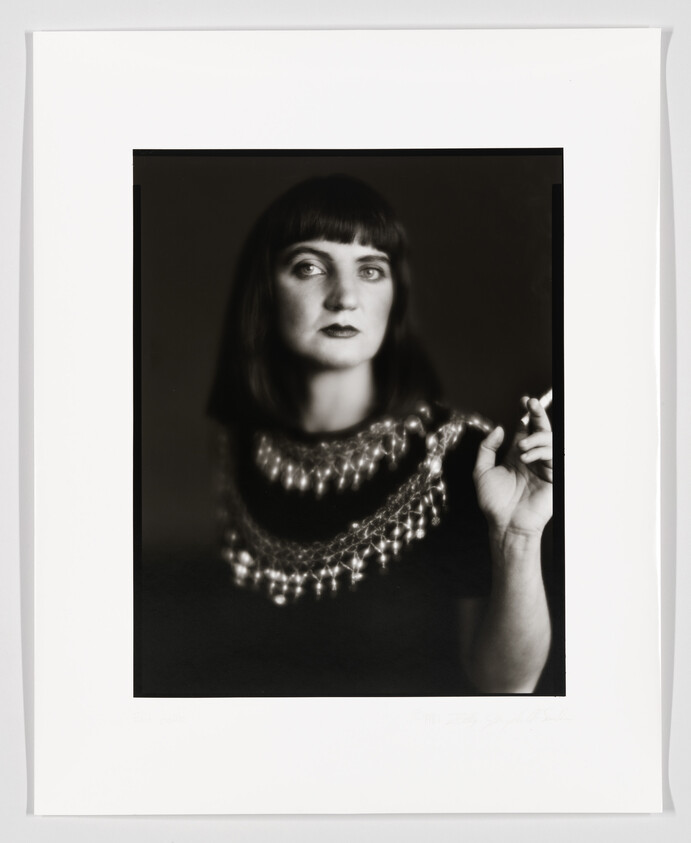 A black and white portrait of a woman with a bob haircut, wearing a dark top and a knitted shawl adorned with pearls. She is looking slightly to the side with a contemplative expression, and her right hand is raised to her shoulder. The photograph is framed with a white border, and there appears to be a signature at the bottom right.