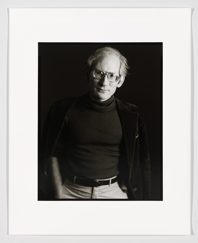 Black and white portrait of a bespectacled man with receding hairline, wearing a turtleneck and jacket, looking directly at the camera. The photo is framed with a white border and signed at the bottom.