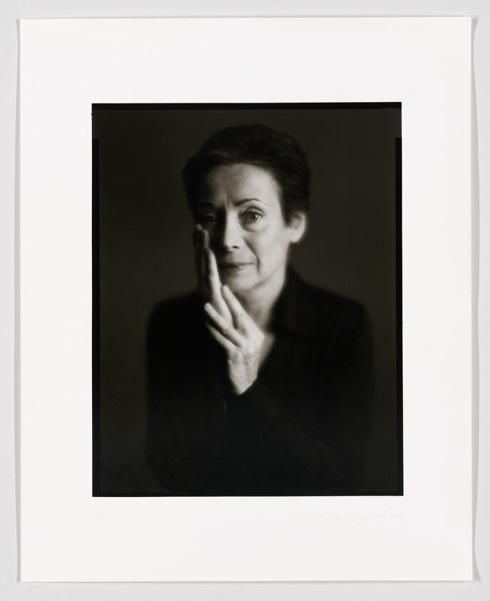 A black and white portrait of a woman with her hands gently pressed together near her cheek, looking contemplatively towards the camera. The photograph is framed with a wide white border, and there appears to be a signature at the bottom.