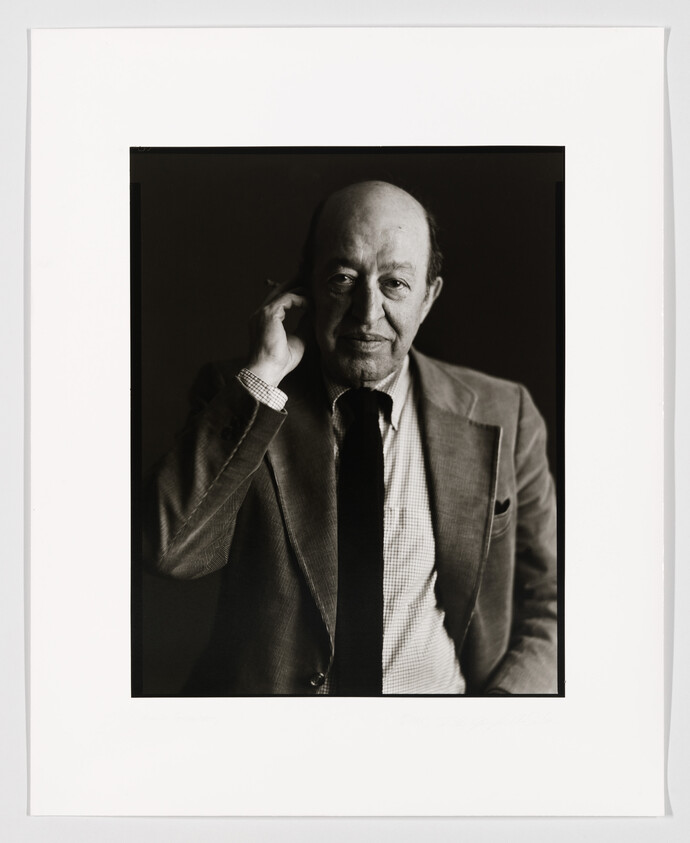 A black and white portrait of a balding man with a confident expression, wearing a suit and tie, and holding his glasses in his right hand. The photo is framed with a white border, and there is a signature at the bottom right corner.