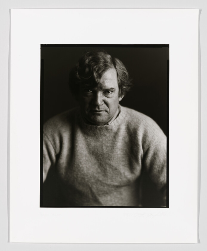 Black and white portrait of a middle-aged man with tousled hair, wearing a crewneck sweater and looking directly at the camera with a serious expression. The photo is framed with a wide white border and signed at the bottom.