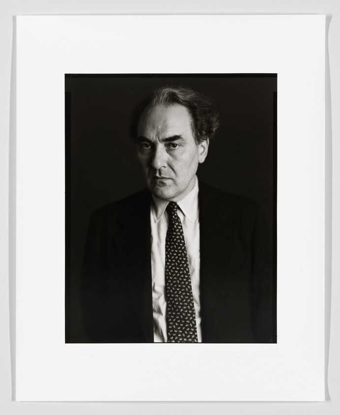 Black and white portrait of a serious-looking man with receding hairline, wearing a dark suit, white shirt, and patterned tie. The photo is framed with a white border that includes a signature and text at the bottom.