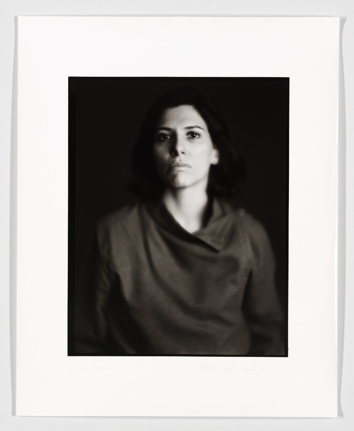 A black and white portrait of a woman with a neutral expression, centered in the frame against a dark background. The photo is slightly out of focus, giving it a soft appearance. The image is bordered by a wide white mat with some text at the bottom.