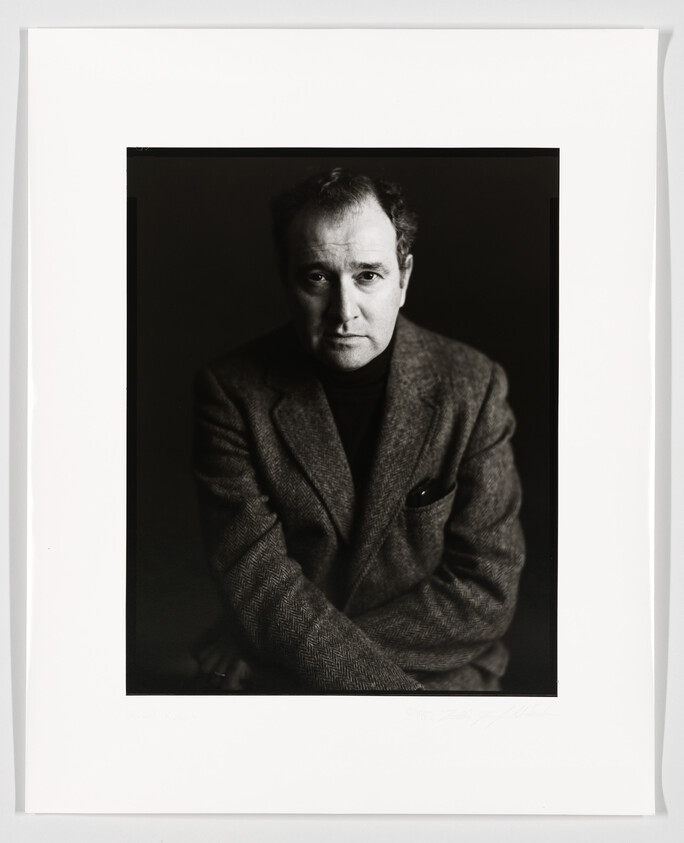 A black and white portrait of a man seated and looking directly at the camera with a serious expression. He is wearing a herringbone jacket and appears to be in a studio setting. The photograph is framed with a white border that includes a signature and some text at the bottom.
