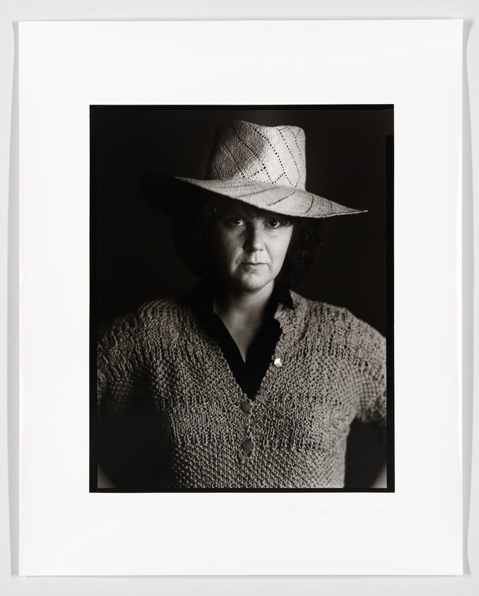 A black and white portrait of a person wearing a textured hat and a knitted sweater, with a focused and serious expression. The image is framed with a white border, and there is a signature at the bottom left corner.