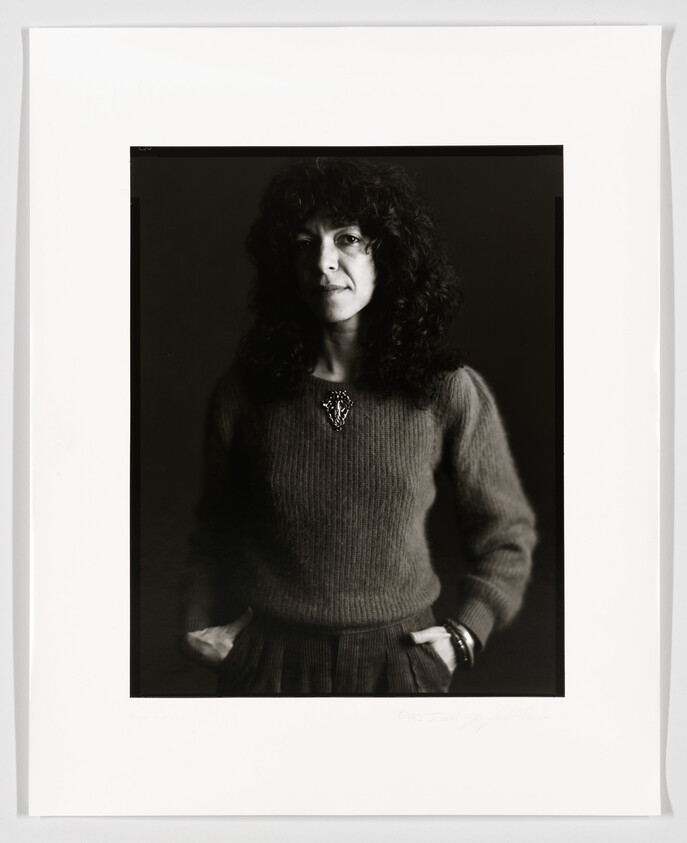 A black and white portrait of a woman with curly hair wearing a sweater and a necklace, hands partially tucked into the pockets of her skirt, with a gentle expression on her face. The photograph is framed with a white border, and there is a signature at the bottom right corner.