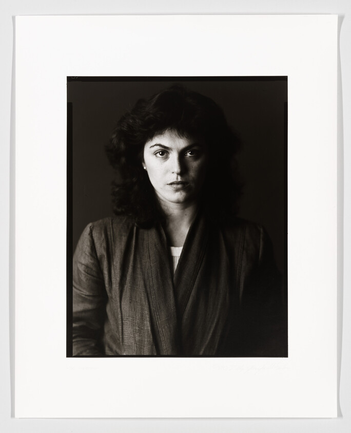 A black and white portrait of a woman with shoulder-length curly hair, looking directly at the camera with a neutral expression. She is wearing a dark blazer over a light shirt. The photo has a white border with a signature at the bottom.