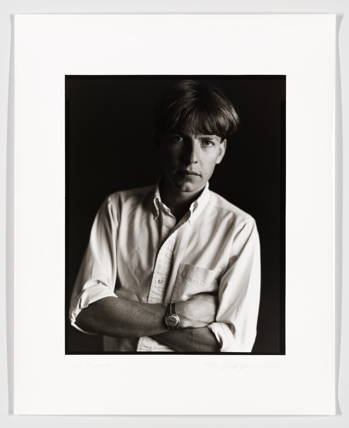 A black and white portrait of a young man with short hair, wearing a white button-up shirt and a watch on his left wrist. His arms are crossed, and he is looking directly at the camera with a serious expression. The image is framed with a white border, and there is a signature and date at the bottom.