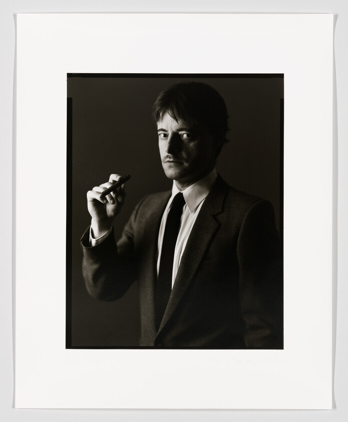 A black and white portrait of a man with a contemplative expression holding a cigar in his right hand. He is wearing a suit with a tie and has a mustache. The photo is framed with a white border, and there appears to be a signature at the bottom right corner.