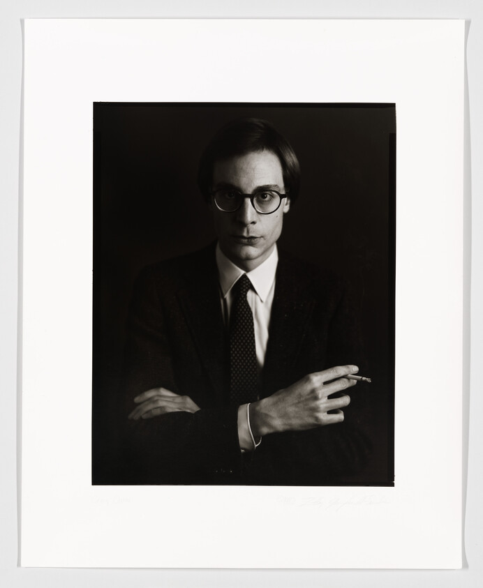 A black and white portrait of a man with round glasses, wearing a dark suit, tie, and a white shirt. He is seated with his arms crossed, holding a cigarette in his right hand. The photo is framed with a white border, and there is a signature at the bottom.