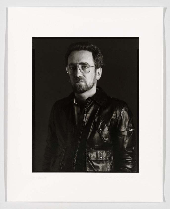 A black and white portrait of a man with glasses and a beard, wearing a leather jacket. The photo is framed with a white border, and there appears to be a signature at the bottom.