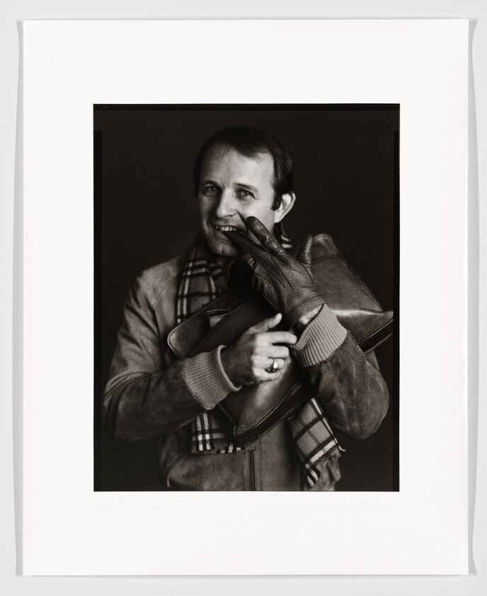 Black and white portrait of a smiling man wearing a plaid scarf and a leather jacket, holding a pair of gloves. The image is framed with a white border that includes a handwritten signature and text at the bottom.
