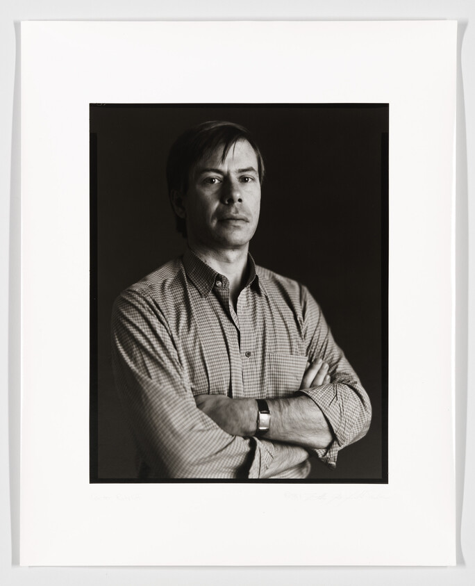 A black and white portrait of a man with his arms crossed, wearing a checkered shirt and a watch, looking directly at the camera. The photo is framed with a white border and has signatures at the bottom.