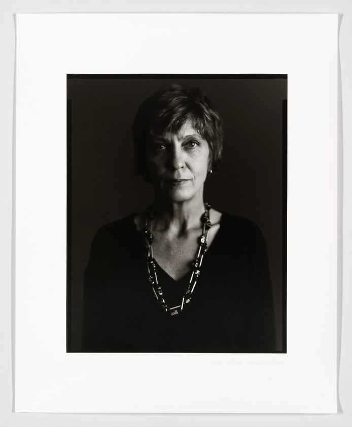 A black and white portrait of a woman with short hair, wearing a dark V-neck top and a beaded necklace, looking directly at the camera with a neutral expression. The photograph is framed with a wide white border, and there appears to be a signature at the bottom right corner.