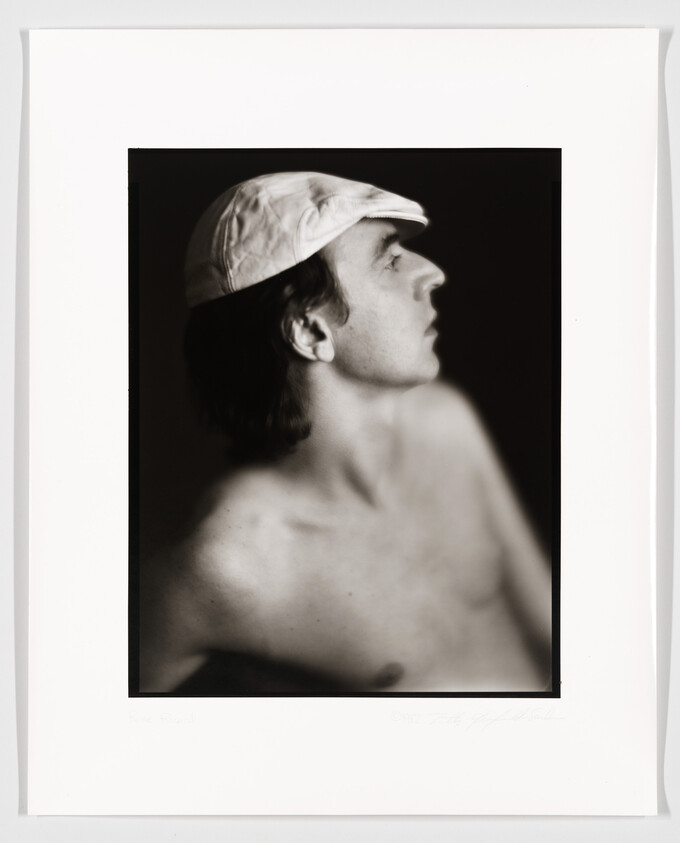 A black and white portrait of a shirtless man in profile wearing a light-colored cap, with a focused and contemplative expression. The photograph is framed with a white border, and there is a signature and date at the bottom right corner.